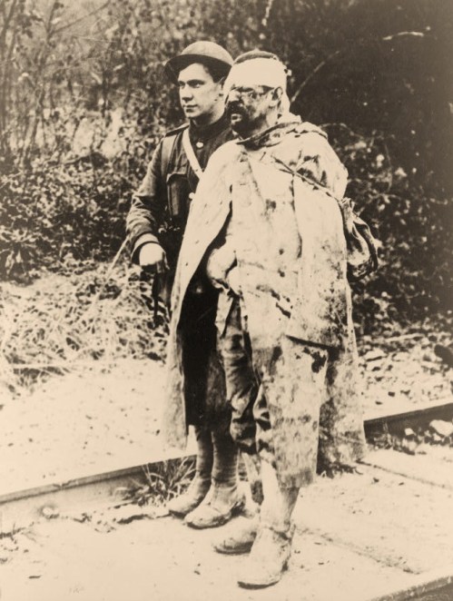 An orderly escorting a wounded, captured soldier to a field hospital for treatment.